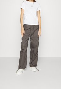 Bílý tričko s krátkým rukávem a logem na hrudi, doplněné volnými tmavě šedými cargo kalhotami se zipovými a knoflíkovými detaily. Bílé obuv.