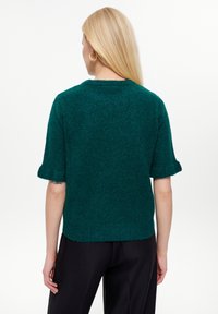 Maglione verde a maniche corte in maglia con scollatura rotonda, tessuto testurizzato e bordo a costine. Indossato con pantaloni neri, che mostrano un look casual.
