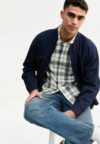 Giacca bomber navy su una camicia a quadri con tonalità di grigio e bianco, abbinata a jeans in denim azzurro chiaro. Tessuto morbido e vestibilità casual.