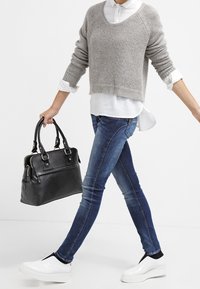 Person som går, klädd i blå jeans, grå tröja över vit skjorta, vita slip-on skor, hållande en svart läderhandväska i höger hand.