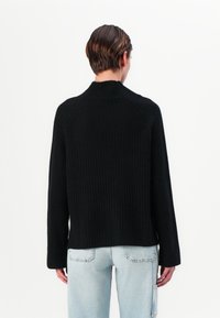 Pull en maille côtelée noir avec un col haut et des manches longues, vu de dos. Associé à un jean bleu clair, mettant en avant une coupe décontractée.