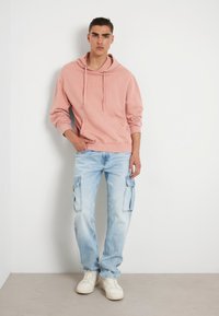 Rausvas su kapišonu džemperis su iškilniu "GUESS" logotipu, derinamas su šviesiai mėlynomis cargo kelnėmis su šoninėmis kišenėmis ir baltais sportbačiais.