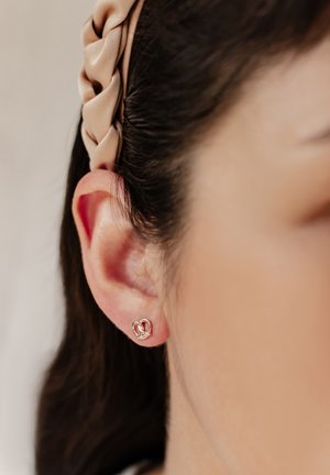 Oreja de mujer con un pequeño pendiente de oro en forma de corazón y una diadema trenzada beige que sujeta el cabello oscuro hacia atrás.