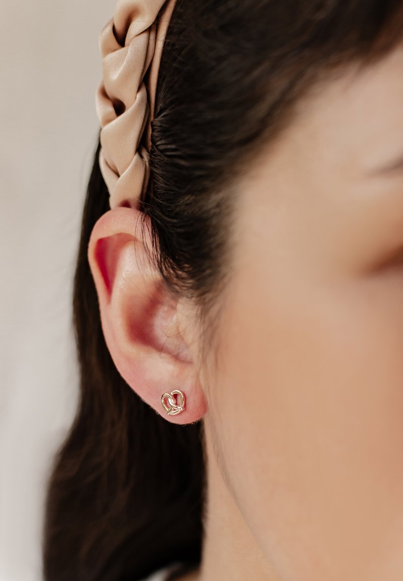 Oreja de mujer con un pequeño pendiente de oro en forma de corazón y una diadema trenzada beige que sujeta el cabello oscuro hacia atrás.