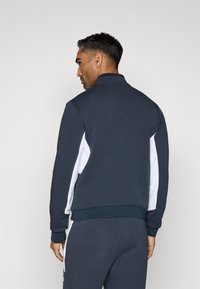 Tmavomodrá fleecová bunda s vysokým golierom a bielymi postrannými panelmi. Obsahuje rebrované manžety a spodný lem, hladkú textúru a atletický strih.