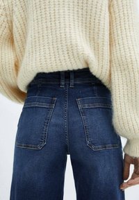 Lichtbeige, textuur gebreide trui gecombineerd met donkerblauwe denim jeans met hoge taille, voorzien van twee achterzakken en contrasterende stiksels.