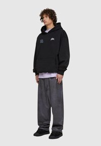 Felpa nera con cappuccio e tasca frontale, caratterizzata da grafiche testuali blu e bianche. Indossata con pantaloni grigi ampi e sneakers nere.