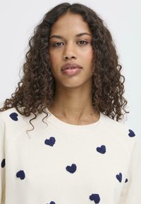 Felpa crema con ricami a forma di cuore blu navy in tutto il capo. Il tessuto ha una texture morbida, con scollatura rotonda e vestibilità rilassata.