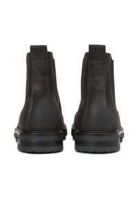 Une paire de bottes Chelsea en suède noir avec des languettes de traction et des semelles en caoutchouc épaisses, présentée de dos sur fond blanc.