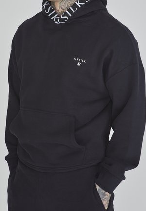 Haut à capuche noir avec une poche frontale, arborant un logo sur la poitrine et une capuche intérieure à motifs. Tissu doux avec une coupe décontractée.