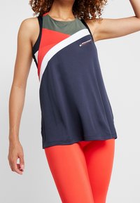 Marinblå idrottstanktop med nätliknande gröna detaljer och diagonala färgblock i röd och vit, tillsammans med klara orangea leggings. Slät tyg.