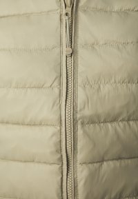 Beige gesteppte Weste mit einem zentralen Reißverschluss und horizontalen Steppnähten. Glatte Textur mit minimalistischem Design. Der Reißverschluss hat einen runden Ziehgriff.