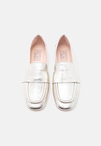 Ζευγάρι λαμπερών ασημί δερμάτινων μοκασίνων με στρογγυλεμένες τετράγωνες μύτες και φαρδιές διακοσμητικές λουρίδες, εμφανιζόμενα από πάνω σε απλό φόντο.