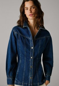 Mørk denimjakke med tettsittende kropp, knappefront og lange puffermer. Har stikninger og sølvfarget maskinvare.