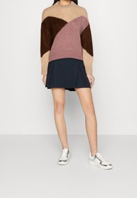 Femme portant un pull color-block beige, marron et rose, une mini-jupe plissée bleu marine et des baskets blanches avec des détails argentés.