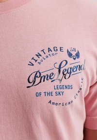 Ružičasta pamučna majica s tamnosivim vintage stilom grafičkog teksta: "Pme Legend", "Legends of the Sky", sa naglascima krila i kružnim dizajnom.