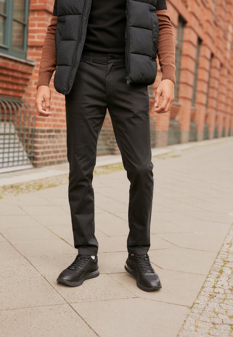 Personne debout sur un trottoir en ville portant un pantalon noir, des baskets noires, un gilet ouatiné noir et un haut à manches longues marron, devant un mur en briques.