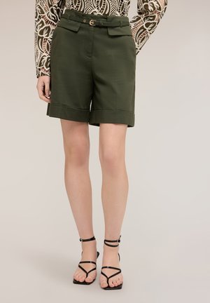 Femme portant un short vert foncé à taille haute avec revers, une fine ceinture et des sandales à talons à fines brides, debout sur un fond uni.