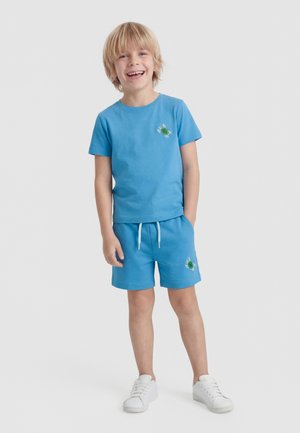 Uśmiechnięty chłopiec o blond włosach, ubrany w niebieski t-shirt i pasujące krótkie spodenki z naszywkami w kształcie żółwi oraz białe sportowe buty, stojący z jedną ręką w kieszeni.