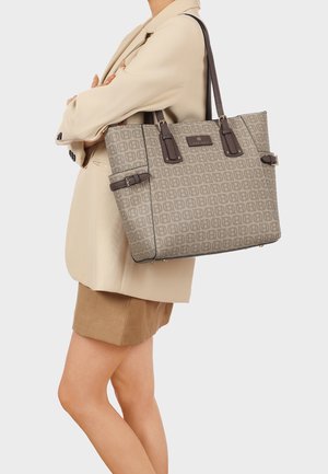 Donna con blazer beige e gonna che porta una grande borsa a mano fantasia con manici marrone scuro sulla spalla.