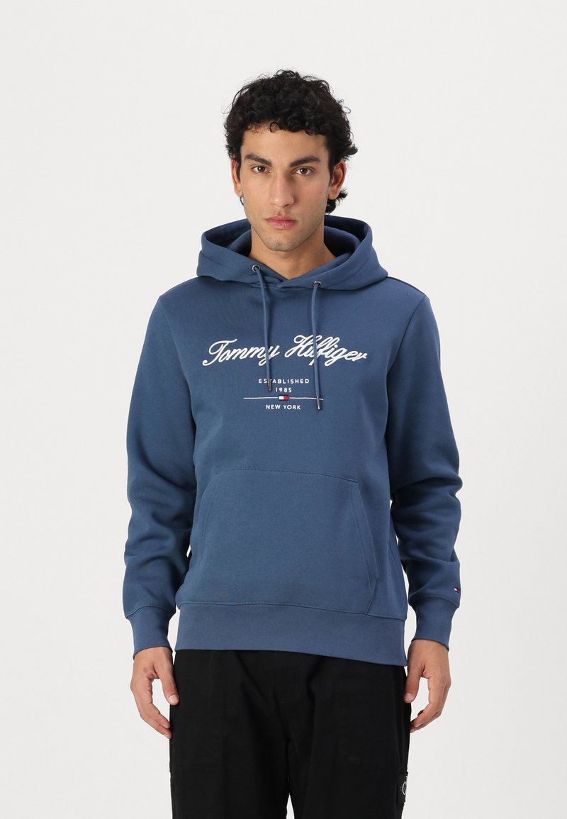 Hanorac albastru din bumbac, cu buzunar frontal, glugă cu șnur și logo-ul "Tommy Hilfiger" în alb. Dispune de manșete și tiv cu nervuri.