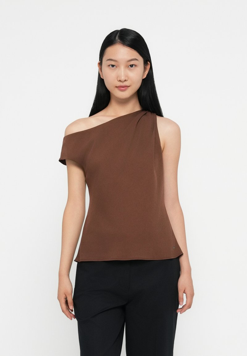 Top brown a spalla unica con tessuto morbido e drappeggiato e una consistenza liscia, abbinato a pantaloni scuri. Design semplice con dettagli minimi.