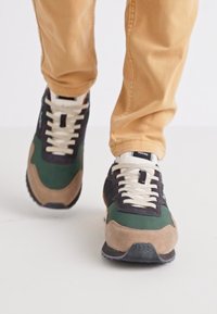 Baskets présentant une combinaison de daim vert, marine et beige, avec des lacets blancs et une semelle en caoutchouc noir texturé.