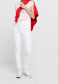 Białe wąskie dżinsy, materiał bawełniany, wysoki stan i pięć kieszeni. Połączone z jasnoróżową koszulką i czerwonym swetrem przewiązanym wokół szyi.