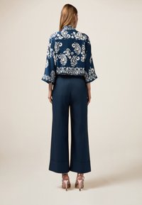 Marineblauwe cropped jas met bloemenpatroon en wijde mouwen, gecombineerd met hoge taille, wijde broek in een donkere tint en metallic hakken met bandjes.