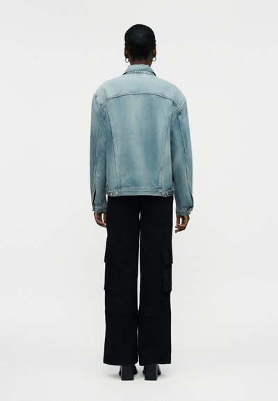 Άτομο με κοντά μαλλιά που φοράει ανοιχτό μπλε τζιν μπουφάν, μαύρο φαρδύ παντελόνι cargo και μαύρα μποτάκια με τακούνι, εμφανιζόμενο από πίσω σε λευκό φόντο.