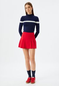 Maglione a collo alto navy con striscia bianca, gonna plissettata rossa, calze al ginocchio navy e sneakers rosse con suola marrone. Tessuto liscio, design aderente.