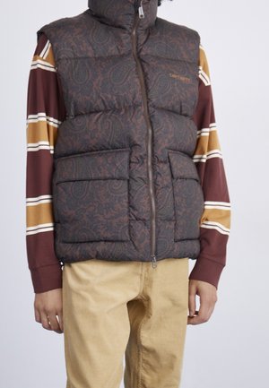 Person trägt eine dunkle, paisleygemusterte Steppweste über einem gestreiften Langarmshirt und beigen Hosen vor einfarbigem Hintergrund.