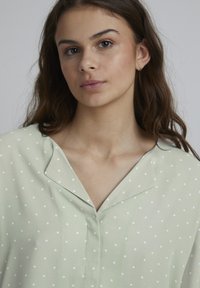 Blouse vert clair à pois blancs, avec un col en V et des manches courtes. Texture douce et coupe ample.