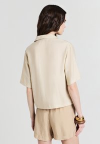 Donna con lunghi capelli castani, che indossa una camicia beige a maniche corte e pantaloni corti abbinati, vista da dietro, con bracciali in legno e oro.