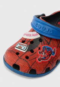 Sabots aux motifs de Spider-Man rouge et noir avec une sangle bleue et trois charms décoratifs Spider-Man à l'avant.