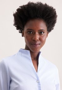 Camicia a righe azzurro chiaro e bianca con colletto alto, dettaglio dei bottoni e texture della stoffa liscia. Primo piano della camicia su un modello.