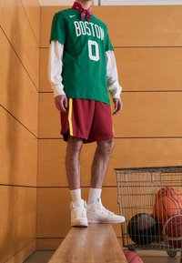 Persoon in een groen Boston-trui, rode shorts, witte sneakers en een rode bandana staat op een gymbank naast een metalen kar met basketballen.