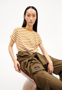 Striped t-shirt in white and orange with short sleeves; paired with loose, olive green pants featuring visible pockets and a branding patch.