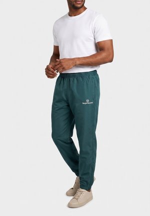 Homme portant un pantalon de survêtement Sergio Tacchini bleu sarcelle foncé, un t-shirt blanc à manches courtes et des baskets beiges, debout devant un fond uni.