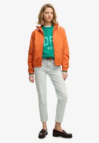 Giacca leggera arancione con cappuccio regolabile, abbinata a una felpa verde, pantaloni di jeans chiari e mocassini scuri.