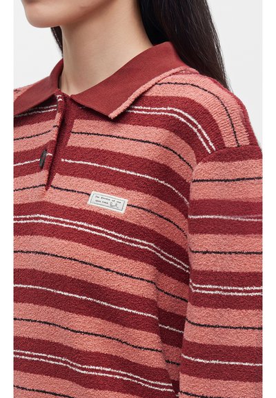 Femme portant un polo tricoté à rayures rouges et roses avec un col et une petite étiquette rectangulaire sur la poitrine.