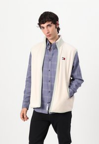 Fleece vest i kremfarge med lys grønn kant, med glidelås foran og sidelommer, brukt over en blå rutete skjorte.