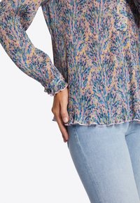 Blusa colorida con pliegues, que combina patrones en tonos rosa, azul y verde. Presenta puños con volantes y un dobladillo redondeado. Se lleva con jeans claros.