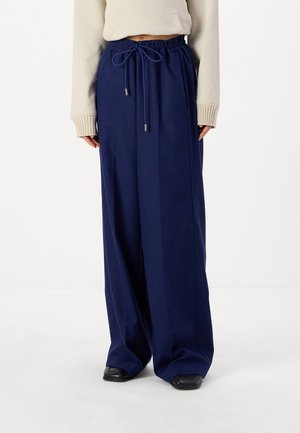 Persoon draagt wijde marineblauwe broek met trekkoord in de taille en zwarte schoenen, gecombineerd met een beige cropped trui.