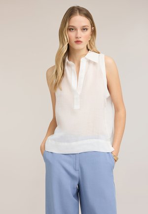 Jonge vrouw met lang blond haar, gekleed in een mouwloze witte blouse en lichtblauwe broek, staand met handen in de zakken tegen een neutrale achtergrond.