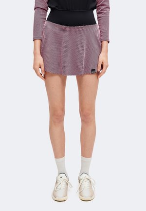 Vrouwelijk model draagt een korte rok met patroon, een zwarte tailleband, witte enkelsokken en witte sneakers, staand tegen een effen achtergrond.