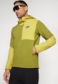 Patagonia kapuce jaku zaļā un dzeltenā krāsā, ar teksturētu audumu, rāvējslēdzēja aizdari un regulējamu kapuci. Iekļautas logotips un sānu kabatas.