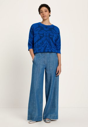 Frau mit kurzen lockigen Haaren, die ein blau gemustertes kurzärmeliges Oberteil, weit geschnittene blaue Jeans und silberne flache Schuhe trägt, steht mit den Händen in den Taschen.