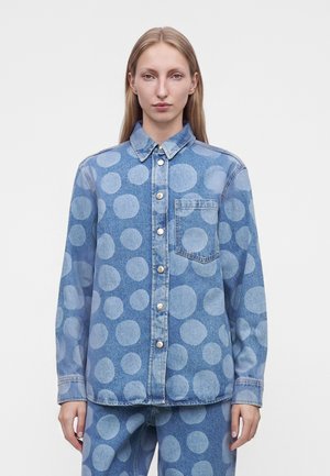 Γυναίκα που φοράει μπλε denim πουκάμισο και παντελόνι με μεγάλα ανοιχτό μπλε πουά, στέκεται μπροστά σε απλό λευκό φόντο.