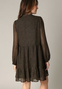 Zwart patroon jurkje met lange mouwen, gerimpelde taille en uitlopende zoom. Gemaakt van doorschijnende, gestructureerde stof met een subtiele glans en geometrisch ontwerp.